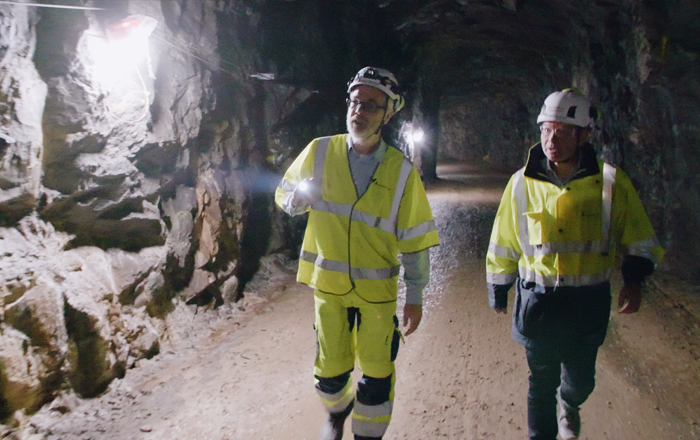 Två personer i arbetskläder. Går på en väg i en tunnel med ficklampa och inspekterar tunnelväggarna..