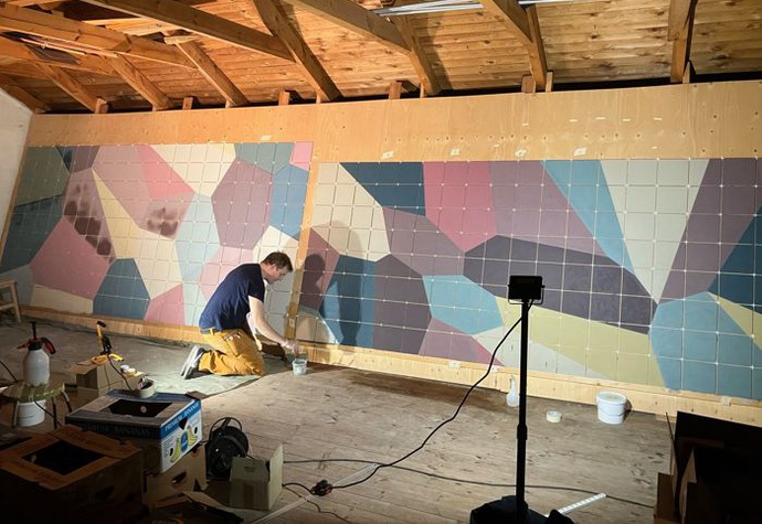 En man sitter på huk och målar framför en väg med geometriska mönster i olika färger.