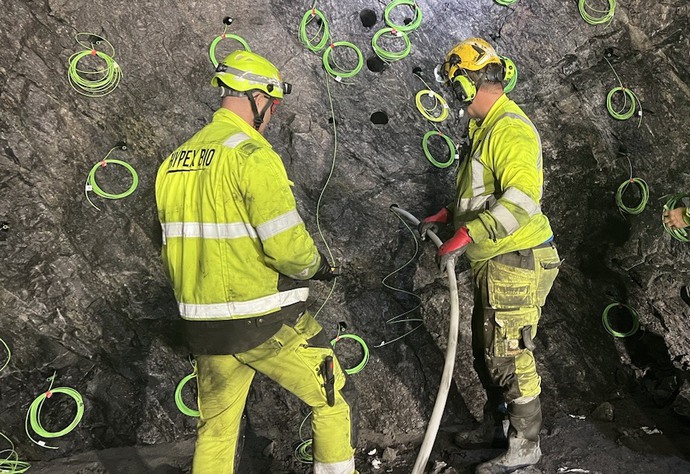Två personer i gula arbetskläder står vid en bergvägg. Berget är fullt av hål, där det sticker ut gröna och gula trådar. En av personerna håller i en slang som går in i ett av hålen.