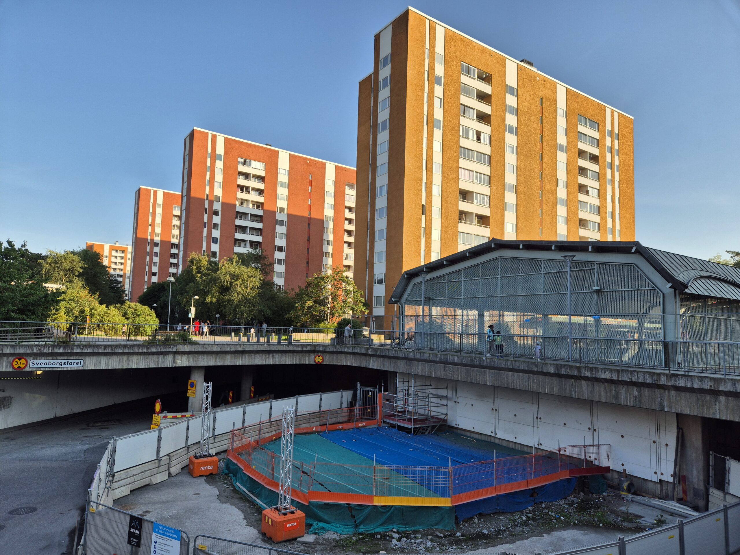 Ett byggarbetsområde inhägnat med plank utanför Akalla station. I bakgrunden går människor. Höga bostadshus finns intill.