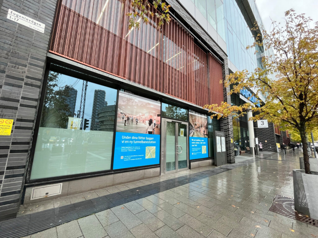 En byggnad med glasfasad och stora skyltfönster med information om tunnelbanestationen Hagastaden som byggs under marken. På trottoaren framför finns en vägskylt, träd med höstlöv och blöta plattor.