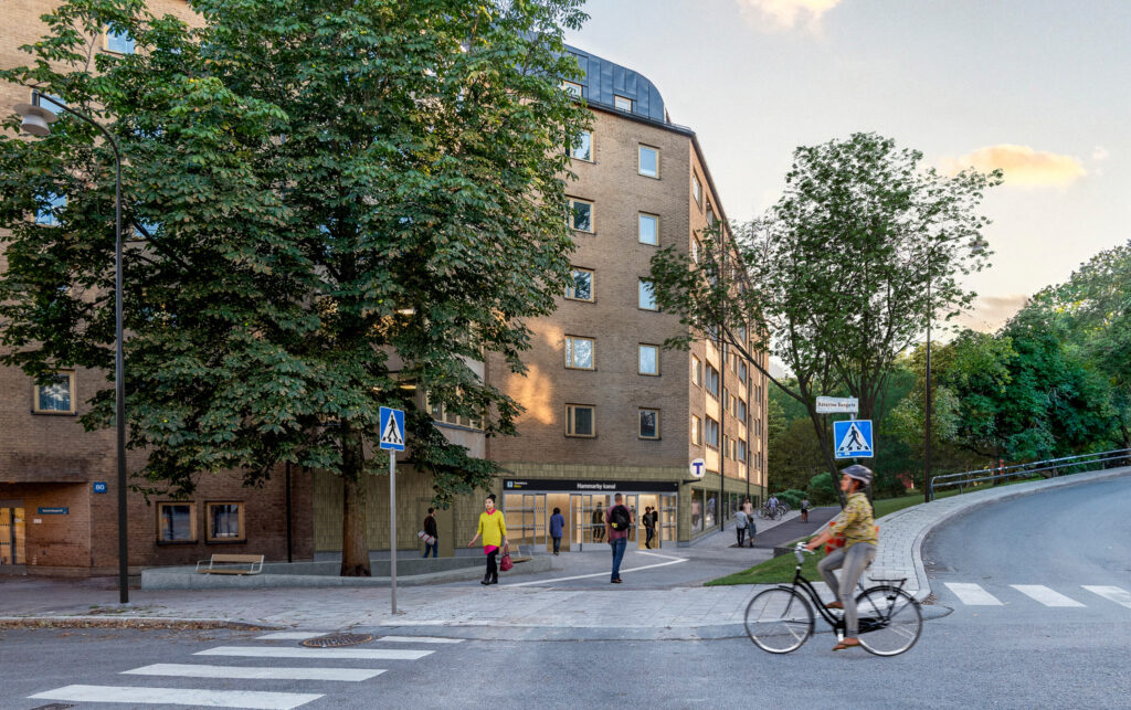 Visionsbild av framtida tunnelbaneentrén vid Hammarby kanal integrerad i en tegelbyggnad, omgiven av träd och fotgängare. En cyklist passerar framför ett övergångsställe i förgrunden.