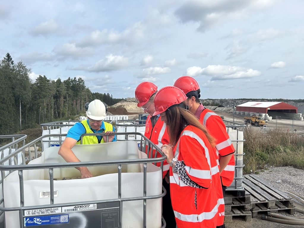 Fyra personer i varselväst och bygghjälm tittar ner i ett stort plastkärl.