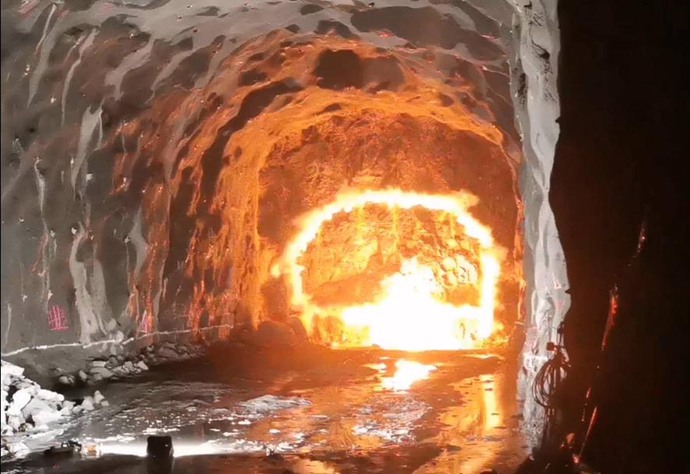 En tunnel under sprängning, där en ljusstark explosion lyser upp den omgivande bergväggen. Rök och värme reflekteras i den blöta tunnelgolvet.