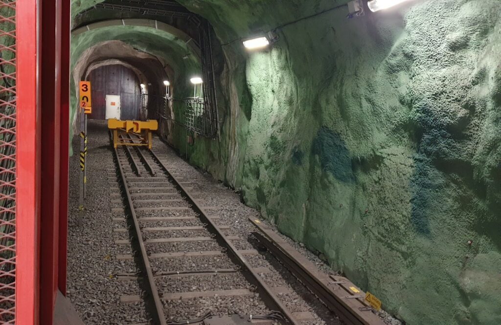 Kungsträdgårdens tunnelbana idag. Ett järnvägsspår slutar vid en buffertstopp i en upplyst tunnel med grönfärgade bergväggar. En dörr syns i slutet av tunneln, och skyltar med siffror är placerade vid spåret.