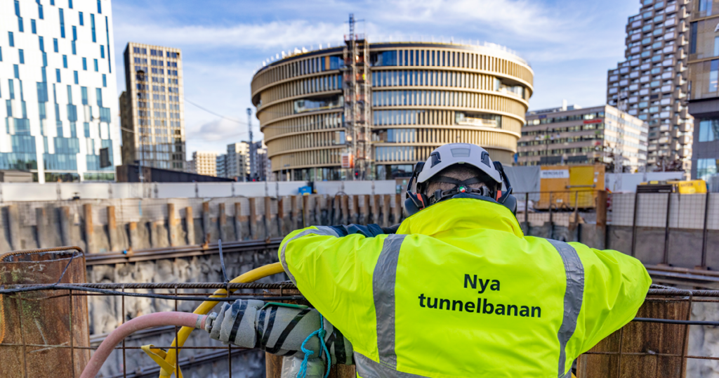 En man i gul jacka med texten nya tunnelbanan på ryggen tittar ut över ett arbetsområde
