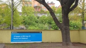 Bilden visar en skylt på det ljusgröna planket runt Stigbergsparken. På skylten står det "Vi bygger ut Blå linje till Nacka och Söderort".