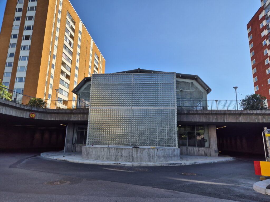 En byggnad med glasfasad framför ett hus. Höga hus i bakgrunden.