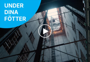 Vy uppåt inuti en djup byggnadsschakt med betongväggar, stålkonstruktioner och ett öppet tak där dagsljus strömmar in. I det övre vänstra hörnet syns en blå cirkel med vit text: "UNDER DINA FÖTTER".