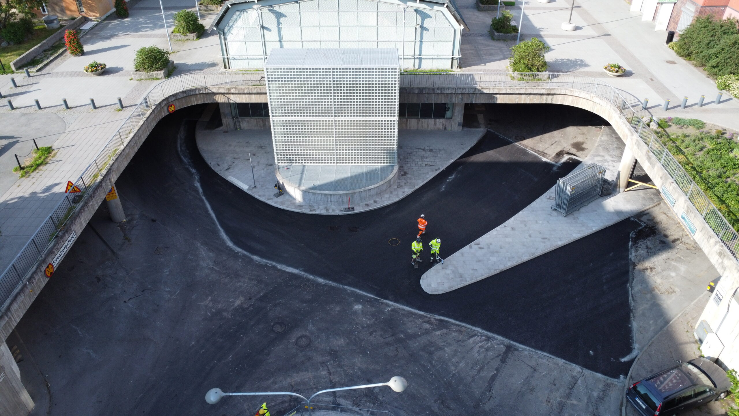 Glasbyggnad på yta med marksten. Asfalterad gata runt om och ovanför finns en större byggnad med glasfasad.