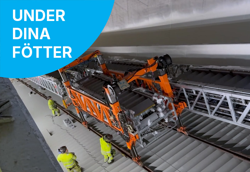 En stor orange maskin arbetar med att bygga en rulltrappa i en tunnelbana, omgiven av flera byggarbetare i varselkläder. Ovanpå bilden syns en blå cirkel med vit text som lyder "UNDER DINA FÖTTER".