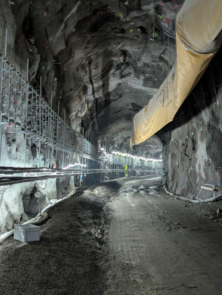 En bergtunnel med arbetsställningar längs ena sidan och en stor ventilationsduk hängande från taket. En person i gul skyddsutrustning står längre in i tunneln.