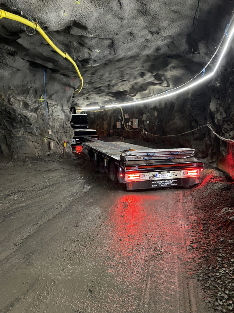 En lastbil med släp kör i en bergtunnel med ojämna väggar och tak. Röda bakljus lyser och en ljusslinga löper längs tunnelväggen.
