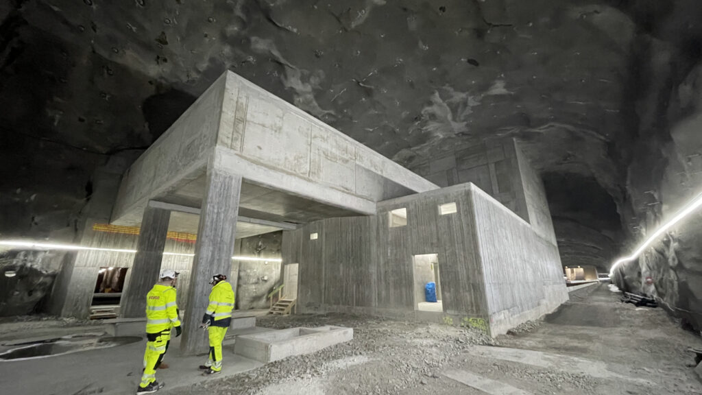 Två personer i gula skyddskläder och hjälmar står i en bergsal framför en stor rå betongkonstruktion. Grova bergväggar och arbetsbelysning omger dem.