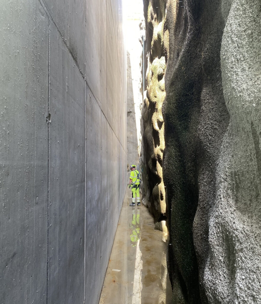 En person i gul skyddsutrustning står i en smal passage mellan en slät betongvägg och en ojämn bergvägg. Marken är blöt och reflekterar ljus.