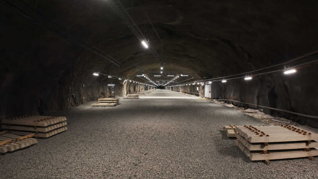 En bred bergtunnel med grusgolv och staplade betongelement längs sidorna. Taket är försett med belysning och installationer.