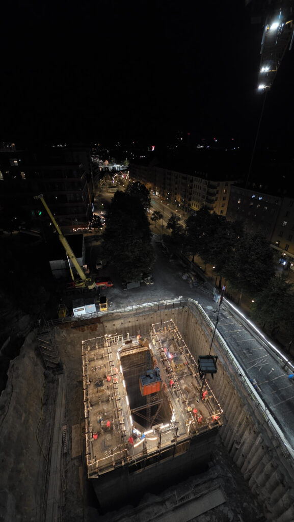 Ett stort upplyst schakt nattetid med byggarbete på botten och flera arbetare i reflexkläder. Ovanför syns stadsgator, träd och byggnader i mörker.