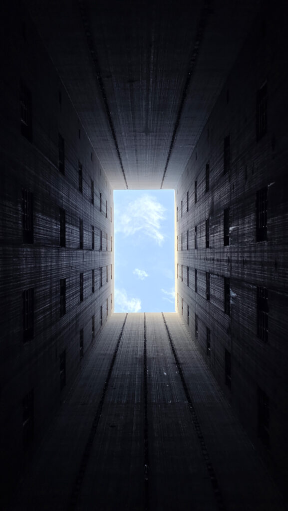 Vy rakt upp mellan fyra höga betongväggar mot en ljus rektangulär öppning med himmel och moln. Små fönster sitter symmetriskt längs väggarna.
