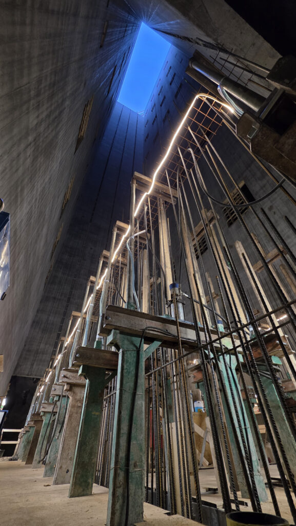 En djup betongschakt med höga väggar sträcker sig upp mot en rektangulär öppning med mörkblå himmel. Längs ena väggen syns byggställningar, rör och arbetsbelysning.