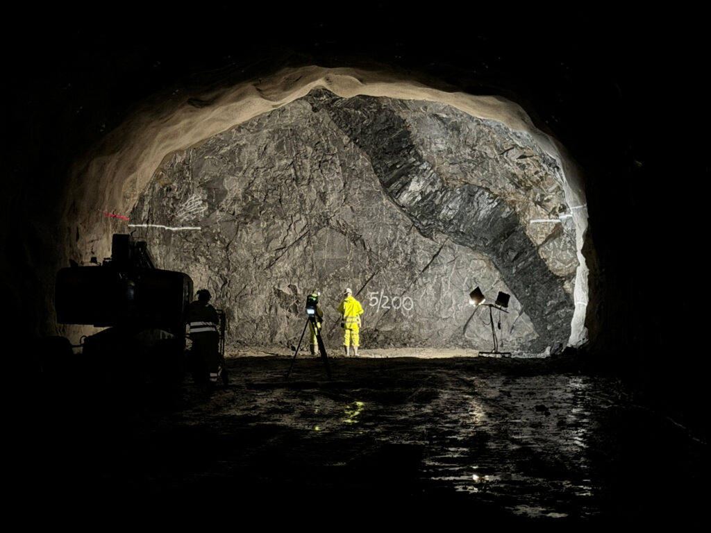 En stor utsprängd bergvägg i en tunnel är upplyst av strålkastare. En person i gul skyddsutrustning står framför väggen och marken är blöt.