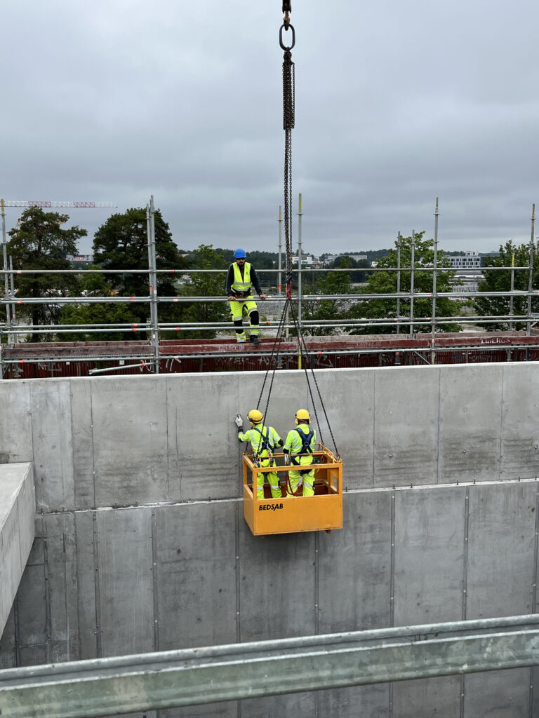 Tre personer i gula skyddskläder arbetar vid en betongvägg, varav två står i en upphängd arbetskorg som lyfts med kran. En tredje person står ovanpå väggen bakom räcke.