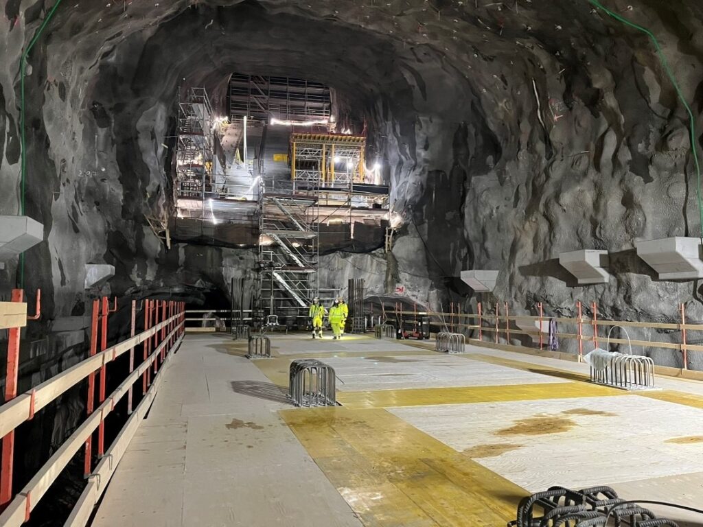 Stor bergtunnel under uppbyggnad med byggnadsställningar, belysning och två personer i varselkläder i mitten. Golvet är täckt av träskivor och stålförstärkningar, och väggarna består av ojämn bergyta med infästa installationer.