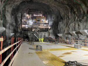 Stor bergtunnel under uppbyggnad med byggnadsställningar, belysning och två personer i varselkläder i mitten. Golvet är täckt av träskivor och stålförstärkningar, och väggarna består av ojämn bergyta med infästa installationer.