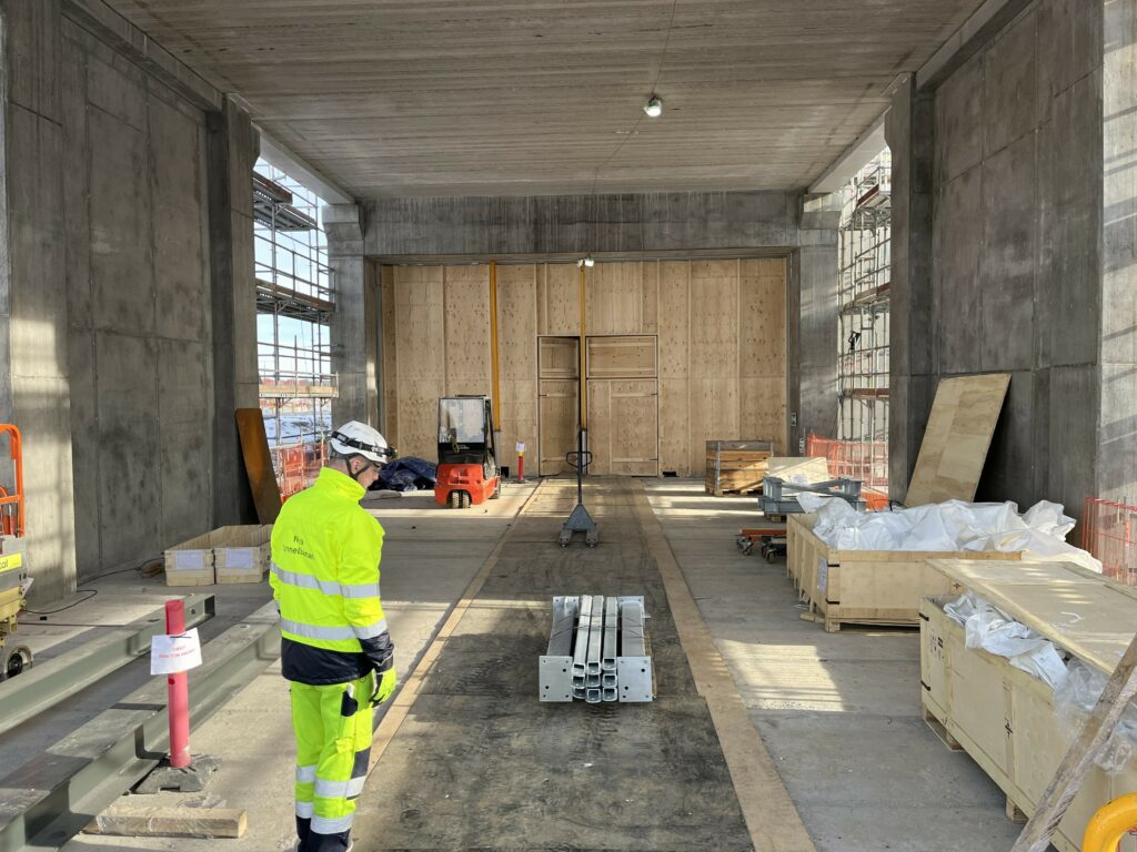 Bilden föreställer ett stort utrymme med betongväggar. Byggmaterial står längs väggar och på golvet. En man i varselkläder står i förgrunden.