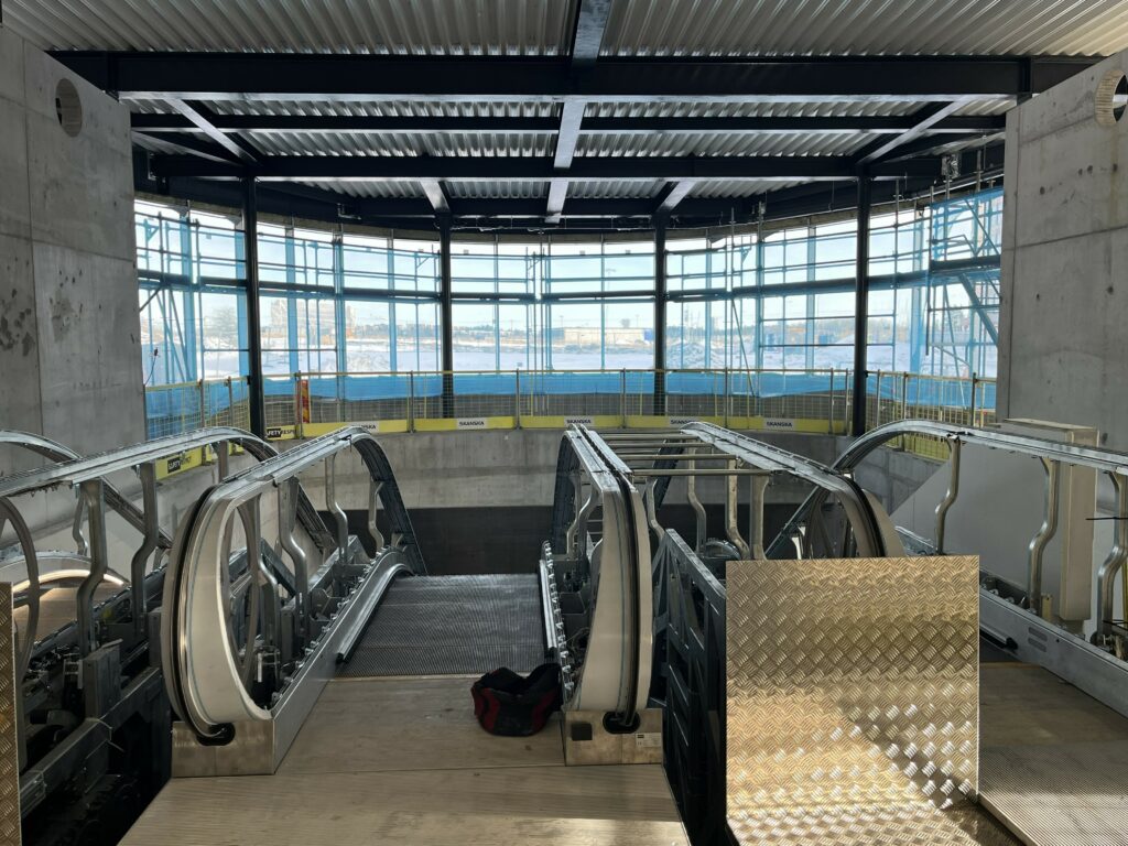 Bilden visar en stor lokal med rulltrappor som leder nedåt. I bakgrunden finns stora glaspartier med byggställningar intill. Arbeten pågår här och där. 