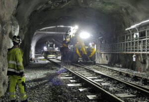 En maskin kör på ett spår inne i en tunnel. En person i varselkläder står vid sidan om spåret och tittar på maskinen.