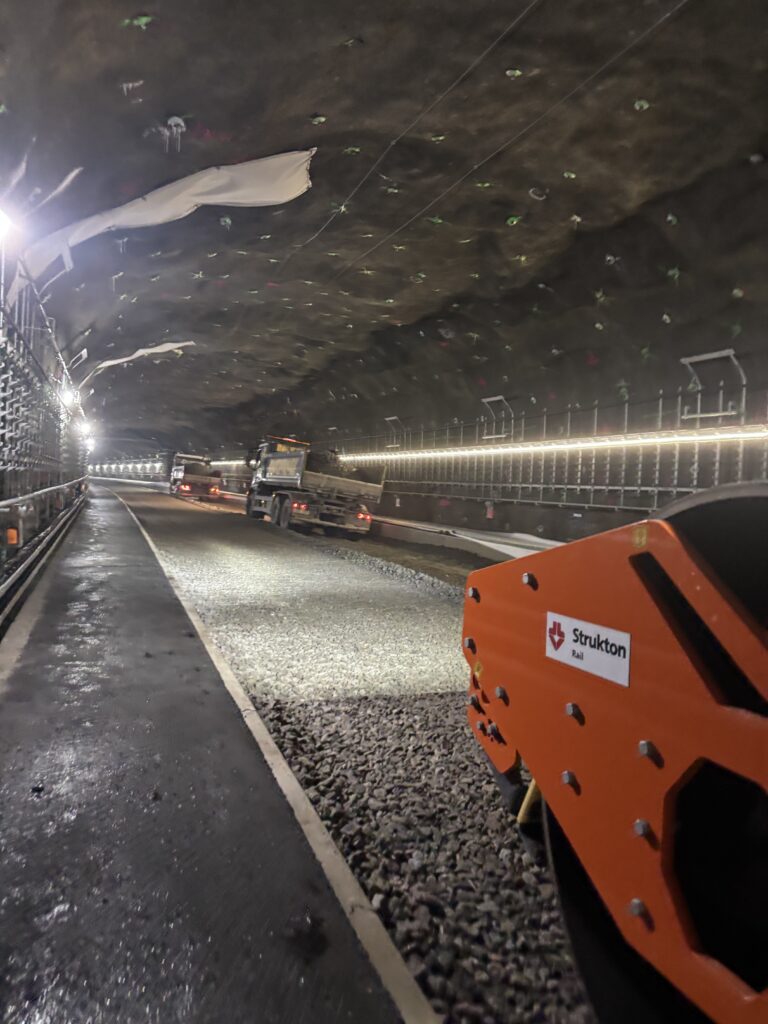 En asfalterad gång bredvid en grusad yta i en tunnel. Två lastbilar och ett annat fordon står på sidan. 