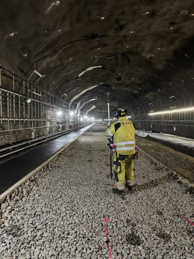 En man med varselkläder står på en grusad yta i en tunnel. Han håller i något avlångt instrument.