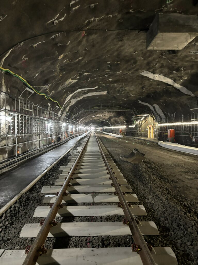 Räls på betongsliprar ligger i en tunnel. Bredvid finns en grusad yta och en asfalterad yta. 