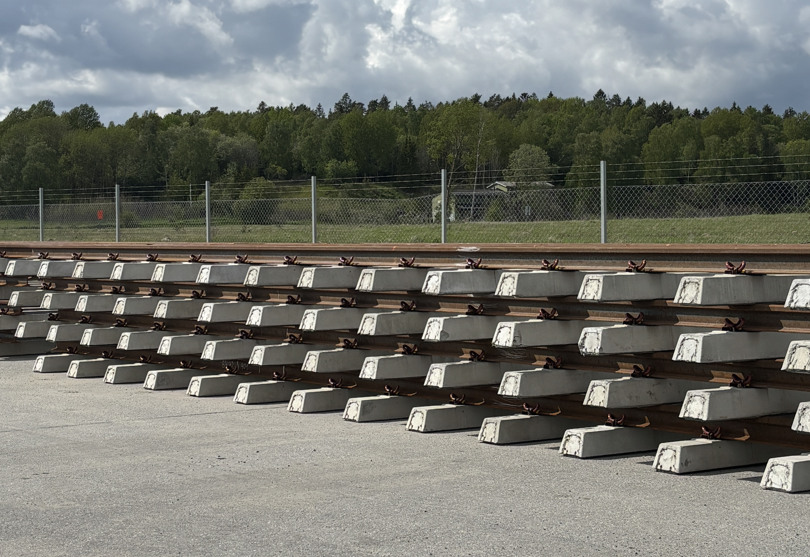 Räls på betongsliprar ligger staplade i flera lager på marken.  