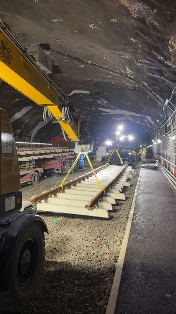 I en tunnel mellan två grävmaskiner hänger en lång sektion med räls på betongsliprar. En man i varselkläder står bredvid en lastbil.