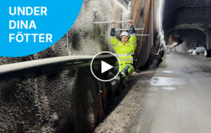 En person i varselkläder och hjälm som hänger med båda händerna i en bult fäst i tunnelväggen, med en tunnelbanetunnel under byggnation synlig i bakgrunden.
